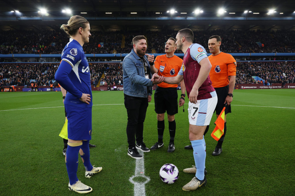 A guest of Budweiser shakes hands with John McGinn of Aston Villa as Referee Craig Pawson speaks with Conor Gallagher of Chelsea at the coin toss p...