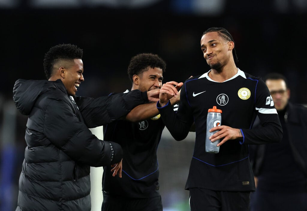 Close-up of Estevao Willian in Chelsea coat, smiling alongside teammates Joao Pedro and Andrey Santos after a match against Napoli.